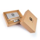 A brown cardboard box holds branded corporate gifts, including a silver power bank, a pen, and a USB drive, all featuring a black logo. The open lid reveals the contents neatly arranged inside.