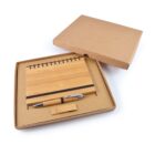A gift set featuring a spiral-bound bamboo notebook, a matching bamboo pen, and a USB drive, all neatly arranged inside an open, eco-friendly cardboard box. The notebook has a black elastic band, and the box is light brown.