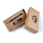 A cardboard gift box containing a black pen and USB drive, both branded with mountain graphics. The box lid, partially open, shows the MOUNTAIN logo. The pen has silver accents, and the USB drive has a small colored label.