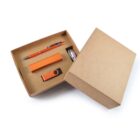 Open cardboard gift box containing an orange pen, an orange USB drive, and a rectangular orange object, possibly a notepad or eraser, arranged neatly inside. The box is partially closed.
