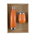 A bright orange insulated bottle and cup set with stainless steel lids, placed inside a plain brown cardboard box. The bottle is taller and cylindrical, while the cup is shorter and rounded.