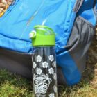 A clear water bottle with a green lid and floral design sits on grass near a blue and gray backpack. The backpack rests on the ground, with visible zippers and exterior pockets. Sunlight casts shadows on the scene, highlighting the outdoor setting.