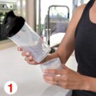 A person in a black tank top is assembling a shaker bottle. They are placing the bottom compartment, filled with powder, onto the main bottle. The kitchen setting features a blurred background. A red number 1 is in the bottom left corner.