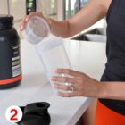 A person in a sleeveless top is scooping protein powder into a clear shaker bottle in a kitchen. A large black container is on the counter. The person has a ring on their left hand. A red number 2 is in the bottom left corner.
