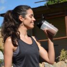 A woman with long dark hair in a ponytail is standing outdoors, smiling while holding a shaker bottle filled with a chocolate-colored drink. She is wearing a black tank top. The background shows a shed and some greenery.
