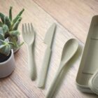 Biodegradable fork, knife, and spoon set placed on a wooden surface next to a small plant in a white pot. A section of a matching biodegradable container is visible on the right.