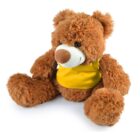 A fluffy brown teddy bear is sitting upright. It has a white snout and a large brown nose, with dark eyes. The bear is wearing a bright yellow bib and has large paws with visible stitching details.