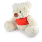 A fluffy teddy bear with cream-colored fur wears a bright red shirt. Its sitting with its legs spread and arms resting on its knees, featuring a friendly smile and a brown nose. The background is plain white.