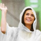 A smiling woman wearing a white rain poncho holds one hand up, enjoying the rain. She looks upward, surrounded by a blurred green background, suggesting the outdoors.