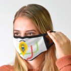 A person adjusts a face mask featuring a subway map design and a Travel Safe logo with a train icon. The mask is white with black edges and colorful lines. The person has long blonde hair and is wearing an orange sweater.