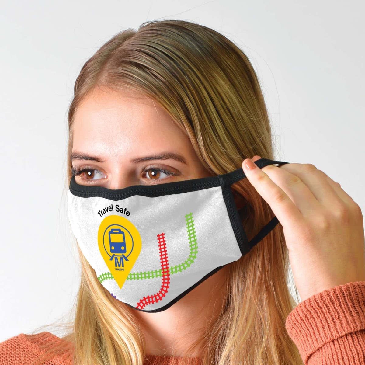 A person adjusts a face mask featuring a subway map design and a Travel Safe logo with a train icon. The mask is white with black edges and colorful lines. The person has long blonde hair and is wearing an orange sweater.