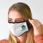 A person with long blonde hair is adjusting a white face mask with black edges. The mask features a green logo and the word ESSENTIAL. The person wears a terracotta-colored garment and has a neutral expression against a light gray background.
