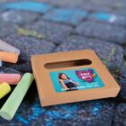 A brown box with a colorful EDUKATE label featuring a smiling girl is on a chalk-decorated pavement. Several colored chalk pieces are scattered nearby. The website www.edukate.com.au is visible on the label.