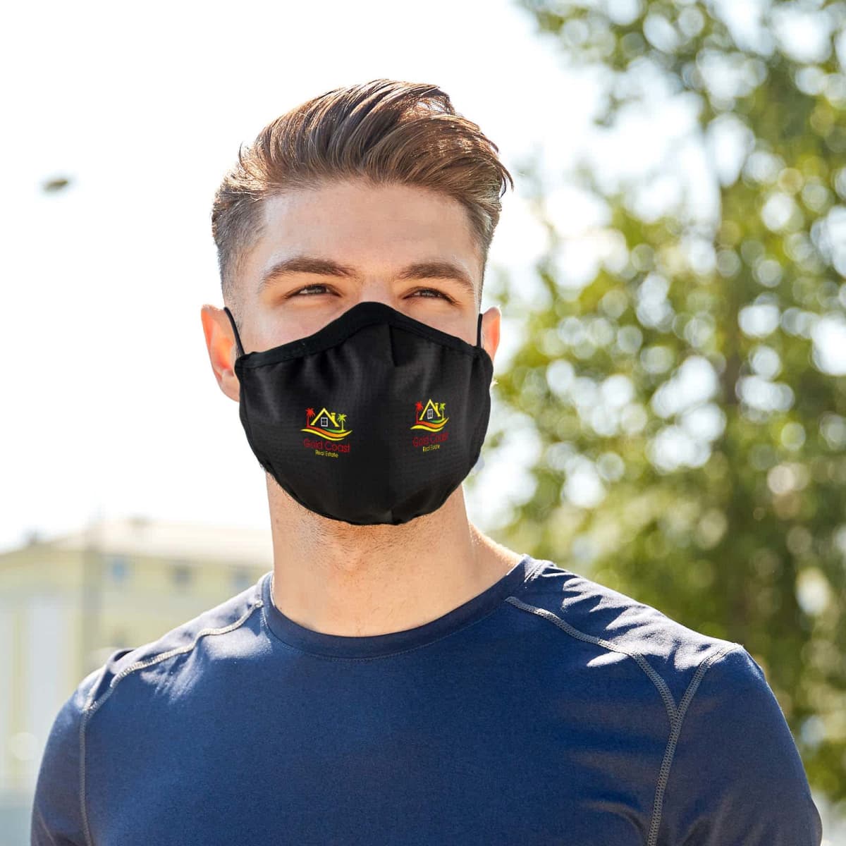 Man with short hair wearing a navy blue shirt and a black face mask standing outdoors. The mask has a colorful design on it. Trees and a building can be seen blurred in the background.