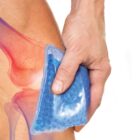 A person holds a blue ice pack on their left knee, which is shown with a red and purple overlay to indicate pain or inflammation. The ice pack is filled with small gel beads. The background is white.