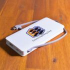 A white power bank with a logo featuring an abstract geometric design and the text E-Xtreme Engineering Group on top. It has a built-in charging cable and is resting on a wooden surface.