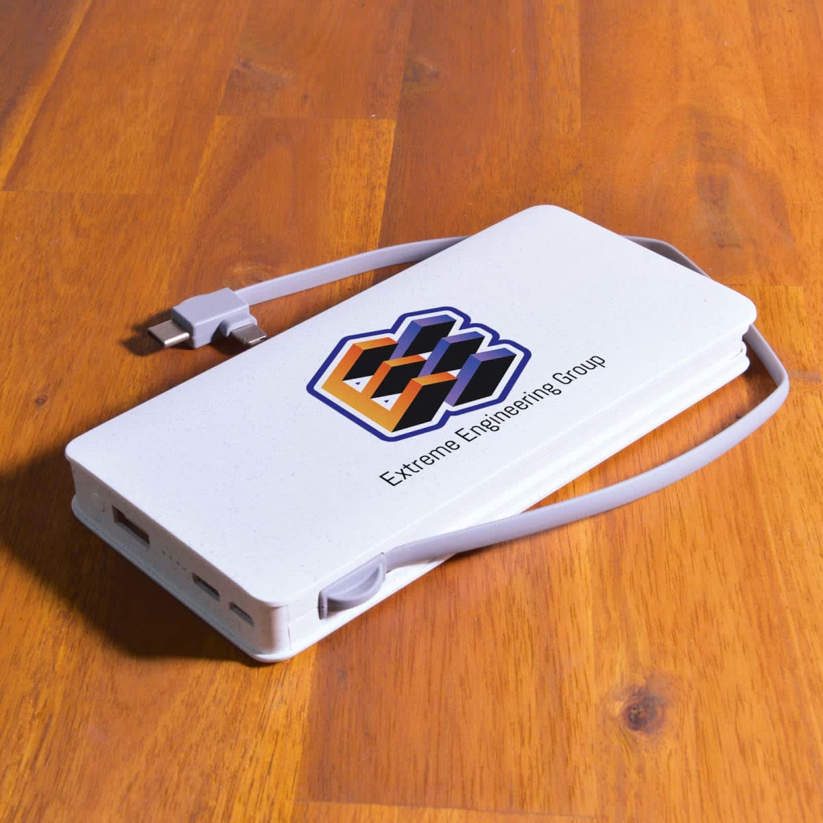 A white power bank with a logo featuring an abstract geometric design and the text E-Xtreme Engineering Group on top. It has a built-in charging cable and is resting on a wooden surface.