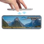 A hand holds a smartphone above a wireless speaker. The speaker displays a scenic mountain view with a lake and grass. Adventure Travel is written on it. Blue waves are shown between the phone and the speaker, indicating connectivity.