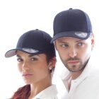 A woman and a man wearing dark baseball caps and white shirts pose side by side against a plain white background. Both caps feature a sticker on the brim. The woman has long hair and earrings, and the man has a beard.