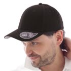 A man wearing a black baseball cap with a NEW MID VISOR sticker on the brim is looking to the side. He has dark hair and a slight beard, dressed in a white shirt.