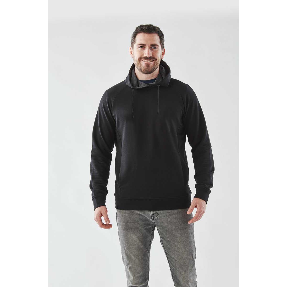 Man wearing a black hoodie and gray jeans, standing and smiling against a white background.