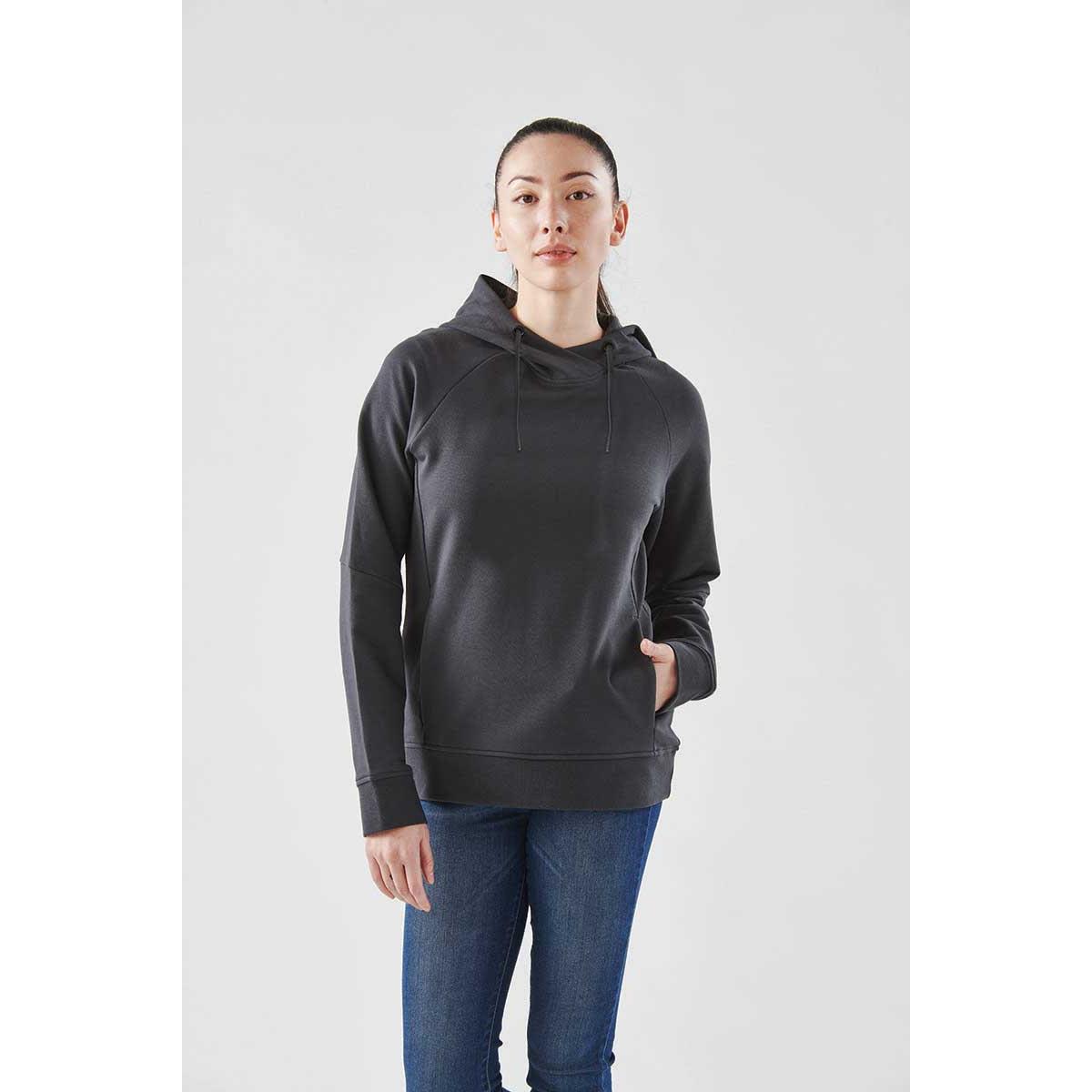 Woman wearing a dark hoodie and blue jeans standing against a plain light background.