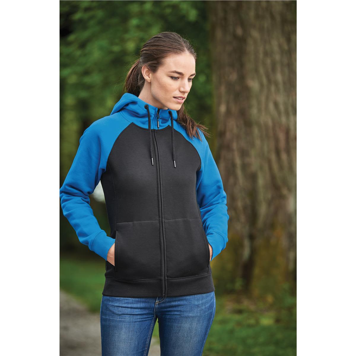 Woman in a black and blue hoodie standing outside with hands in pockets, looking down.