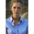 Woman in a blue collared shirt with windblown hair, looking directly at the camera.