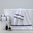 A white bathtub adorned with white towels featuring gray stripes. Two folded towels are stacked nearby on a platform. In front, a transparent tray holds a black soap dispenser and a diffuser with reeds. The background is a gray wall.