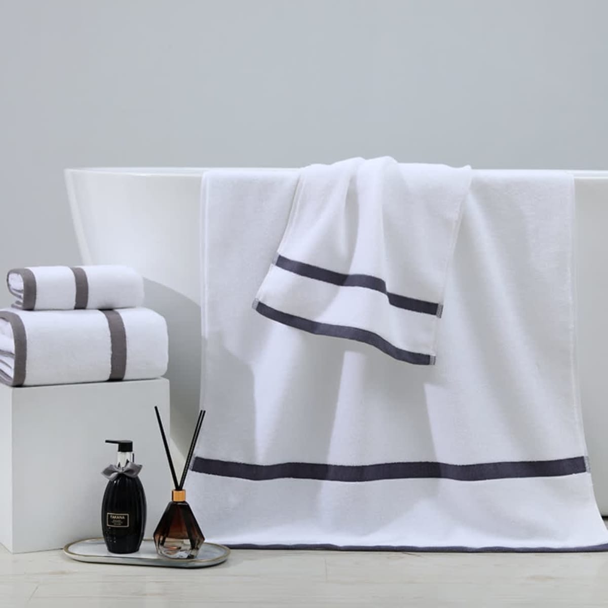 A white bathtub adorned with white towels featuring gray stripes. Two folded towels are stacked nearby on a platform. In front, a transparent tray holds a black soap dispenser and a diffuser with reeds. The background is a gray wall.