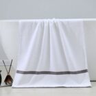 A white towel with a gray stripe near the bottom hangs over the side of a white bathtub. The bathroom features a light gray wall and beige floor. A fragrance diffuser with black reeds stands nearby.