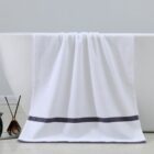A white towel with a dark gray stripe near the edge hangs over the side of a white bathtub. Nearby, there is a dark glass diffuser with reeds on the floor. The background is a light gray wall.