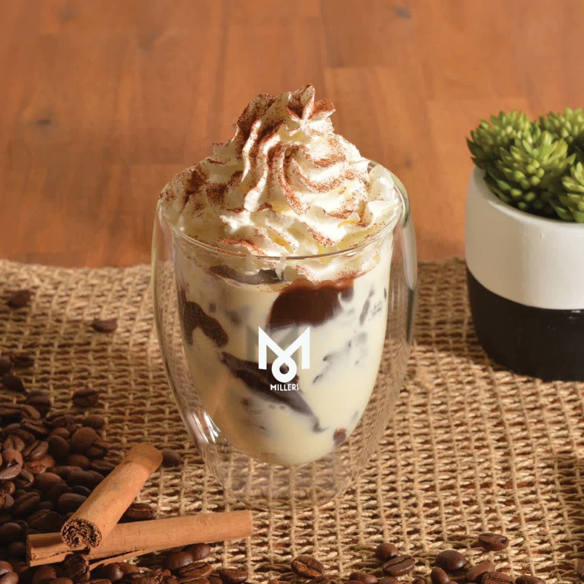 Glass of whipped cream coffee with cinnamon sticks and coffee beans on a woven mat.
