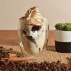 Creamy dessert in a glass with whipped cream and chocolate, surrounded by coffee beans and a plant.