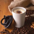 A cup of coffee with froth, surrounded by coffee beans and a lid beside it.