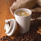Reusable cup of latte with heart art, surrounded by coffee beans on a wooden surface.