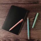 Notebook with a pink pen on it, beside two teal pens on a wooden surface.