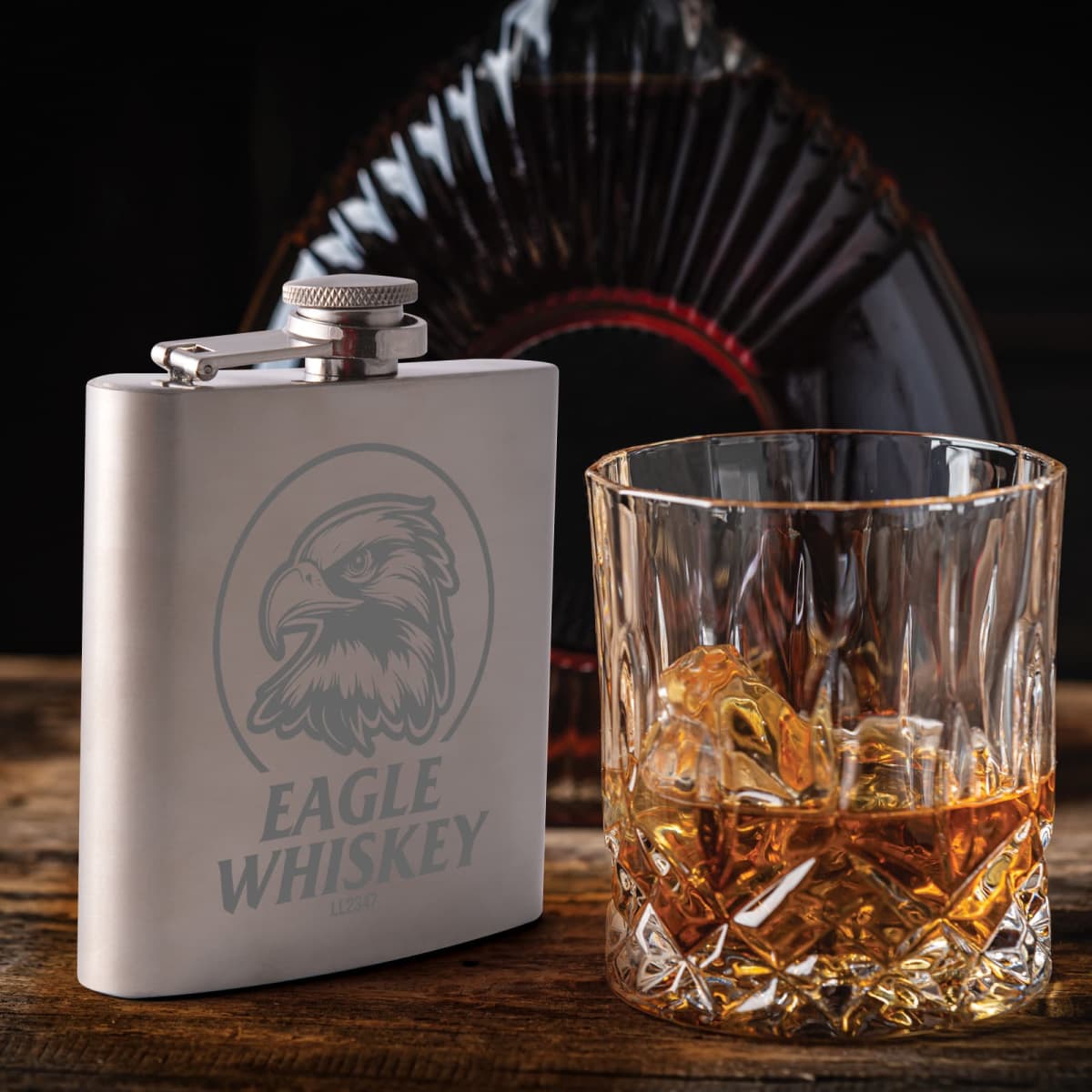 Silver flask with eagle design and Eagle Whiskey text next to glass with whiskey and ice on wooden surface.