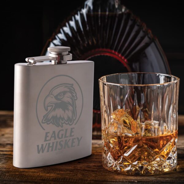 A metal flask and a glass of whiskey with ice on a wooden surface.