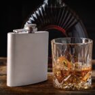 A metal flask and a whiskey glass with ice on a wooden surface.