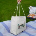 Hand holding a Sprout Community Gardens tote bag on a picnic blanket with snacks nearby.