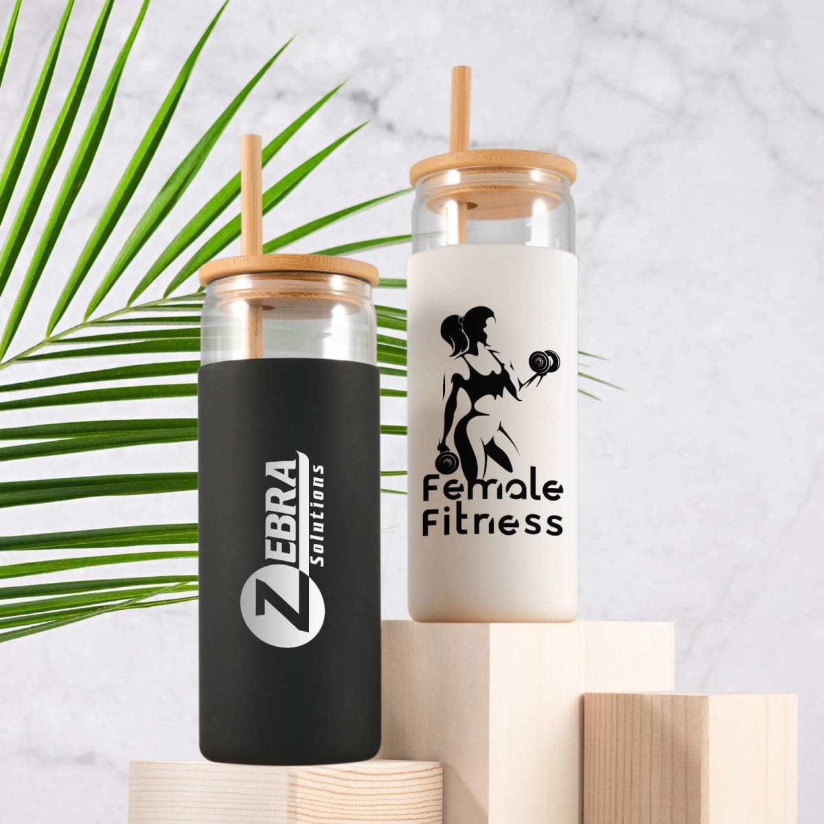Two branded glass bottles with bamboo lids and straws, on wooden blocks. A green leaf in the background.