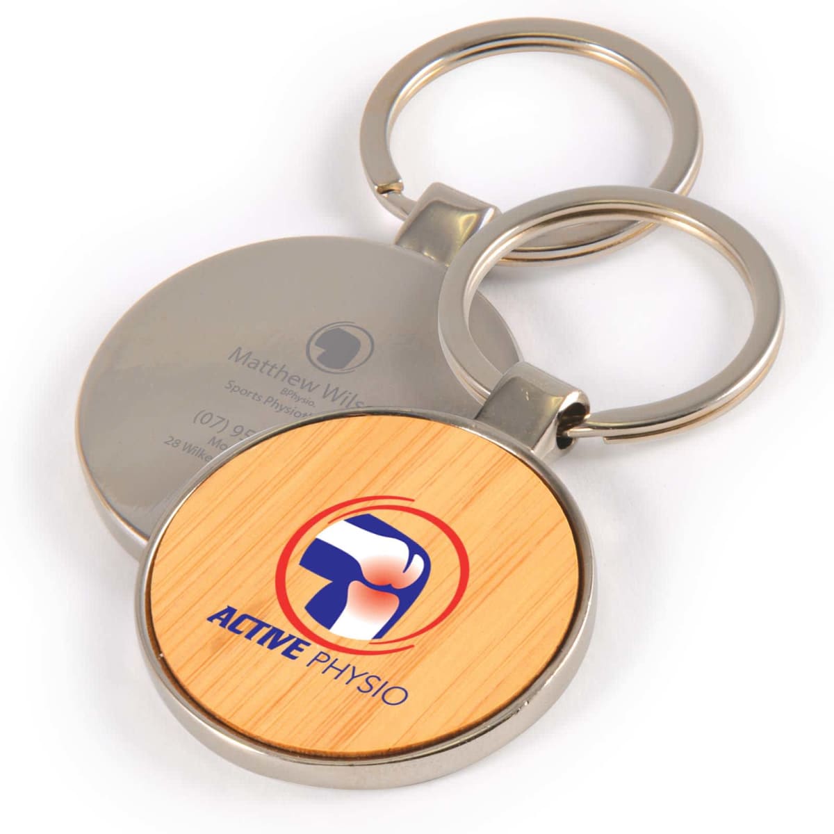 Round metal keychain with wooden center displaying ACTIVE PHYSIO logo; background shows contact info.