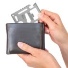 Hands holding a black wallet, inserting a metal multitool card with cut-out features and a ruler on top edge.