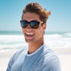 Smiling person in sunglasses and a striped shirt at a sunny beach with waves in the background.