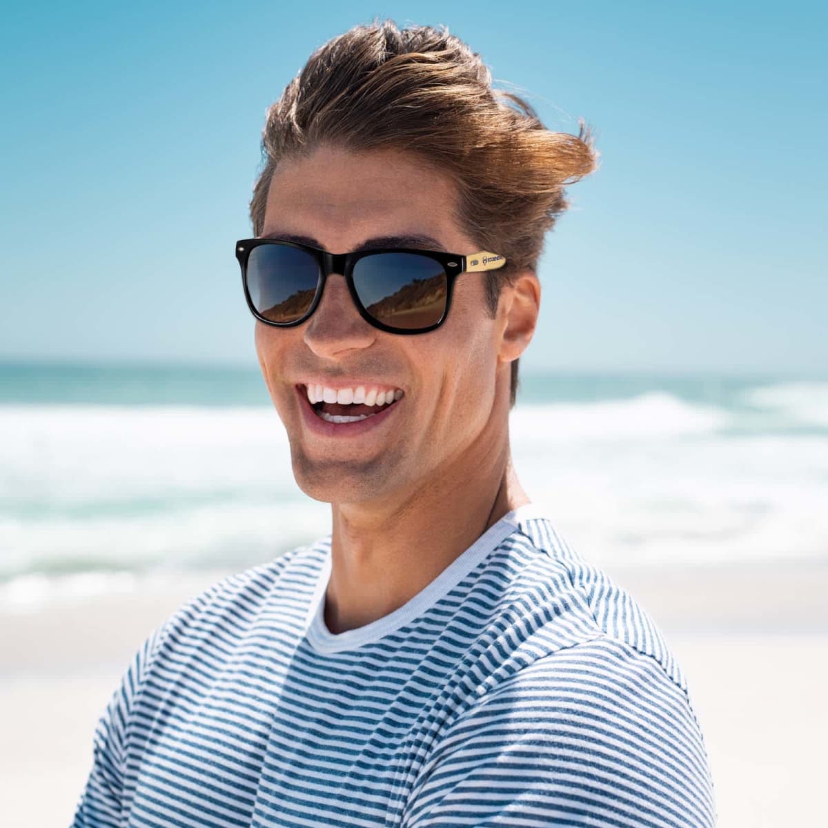 Smiling person in sunglasses and a striped shirt at a sunny beach with waves in the background.