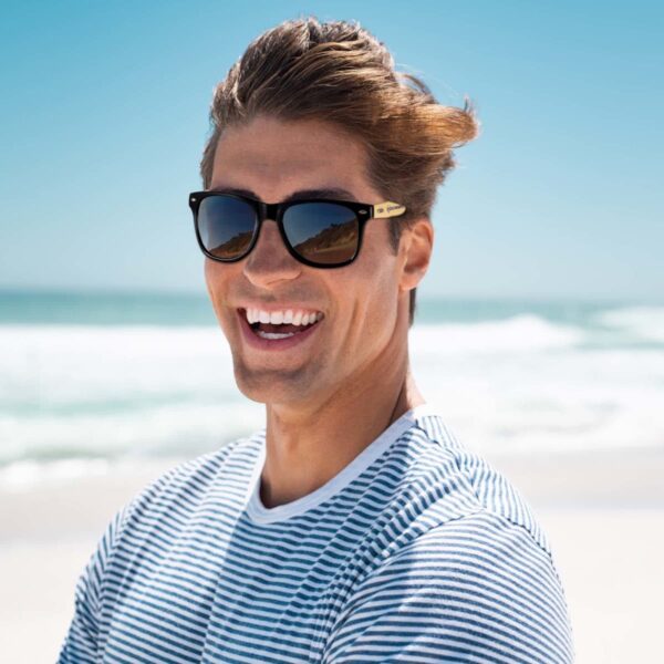 Man with sunglasses smiling at beach, wearing striped shirt, with ocean and clear sky background.