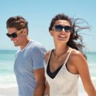 A smiling couple wearing sunglasses walks along a sunny beach with a clear blue sky.