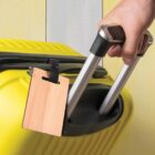Hand gripping handle of a yellow suitcase with a wooden luggage tag attached.