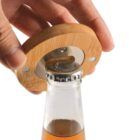 A hand uses a wooden bottle opener with metal components to open a glass bottle with a metal cap.